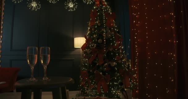 装飾されたクリスマスツリーの近くのテーブルの上に立っているシャンパンの2つの眼鏡を屋内に閉じます 美しいスタジオで新年を祝う人々 新年とクリスマスの概念 — ストック動画