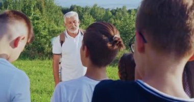Sırt çantası giymiş, çocukların önünde duran, konuşan, açıklama yapan yaşlı bir adamın ön görüntüsü. Çocuklar bakıyor, dinliyor. Doğanın birlikte keşfettiği kavram..