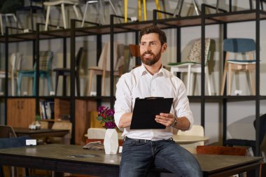 Mobilya mağazasında çalışırken masaya yaslanan mutlu sakallı yönetici. Düşünceli satıcının portresi notlar alıyor, uzaklığa bakıyor, arka planda çeşitli sandalyeler olan raflara karşı. Çalışma konsepti.