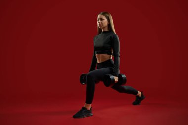 Çömelme hareketleri yapan güzel bir bayan antrenör, kırmızı stüdyoda dambıllarla bacakları büküyor. Güzel enerji ve sportif kadın spor malzemeleriyle egzersiz yapıyor. Egzersiz kavramı.
