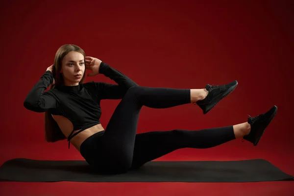 Pretty brunette woman trainer doing twists and abs sitting on yoga mat. Female in black sports suite training cross fit exercise on red studio background. Concept of cardio training.