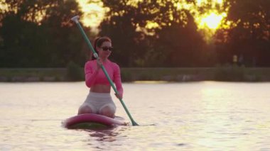 Güneş gözlüklü ve aktif giysili mutlu beyaz kadın şehir gölünde spor yapmak için renkli yiyecek panosunu kullanıyor. Arka planda güzel bir yaz günbatımı. Boş zaman aktivitesi.