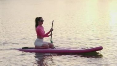 Gün batımında gölde kürek çeken güneş gözlüklü ve spor elbiseli güzel bir kadın. Su sporu ve aktif yaşam tarzları konsepti. 