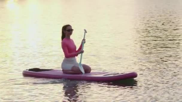 在湖上令人惊奇的落日中 一个体形健壮的年轻女子在桨板上游泳 白种人黑发长桨划船 水上运动的闲暇时间 — 图库视频影像