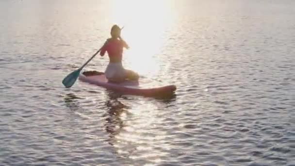 若い健康な女性は 長いパドルでボード上で泳いで 湖の上に素晴らしい夕日を楽しんでスポーツ服を着ています 健康的で活動的なライフスタイルの概念 — ストック動画