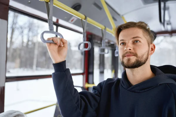 Front view of young male with beard going to work by public transport, standing on bus, waiting. Brunette man holding handle, wearing black khudi. Concept of everyday routine.