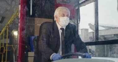 Front view of driver sitting on bus, wearing mask and medical gloves. Old male wih grey hair driving public transport during global pandemic, Concept of pandemic measure.