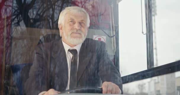 灰色の髪に座っているバスの運転手のフロントビューでは 楽しみにしています 舵を握っている老人は スーツを着て 夢を見て 親切で 仕事を楽しんでいます 都市生活の概念 — ストック動画