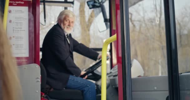 从侧面看公共汽车司机向前望去 乘客们向他致意 头发灰白的老人向戴耳机的女孩和挥手的小男孩打招呼 公共交通的概念 — 图库视频影像