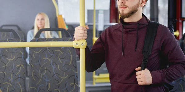 Close up of young man with beard wearing khudi, standing on bus, holding backpack, looking forward. Adult brunette traveling by public transport, busy. Concept of urban life.