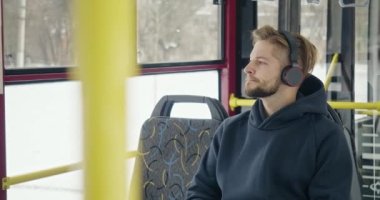Side view of boy with beard traveling by bus. Handsome young man sitting on bus, listening to music in earphones, shaking head. Concept of modern day reality and urban life.