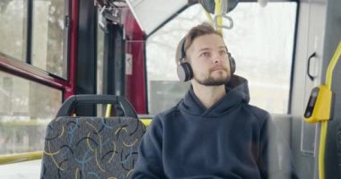 Front view of boy listening to music in earphones on bus. Young man with beard sitting, shaking head, drums playing, looking around, laughing, music enjoying. Concept of youth.