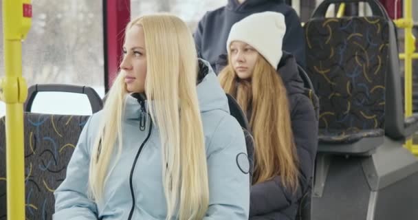 乘客坐在公共汽车上的日常活动的侧视图 穿着蓝色外套的金发女孩笑着 摸着头发 戴着白帽的女孩望着窗户 城市生活的概念 — 图库视频影像