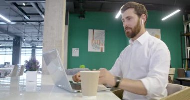 Sakallı esmer adamın laptopta oturduğu, çalıştığı, dizüstü bilgisayarını, kulaklığını, web kamerasını kullandığı yan görüntüler. Yakışıklı satıcı, beyaz gömlek giyen tasarımcı. Kentsel yaşam tarzı kavramı.