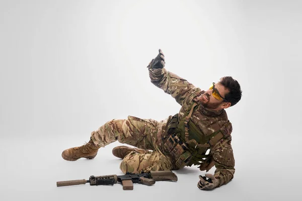 Weak fallen soldier in camouflage asking for help indoors. Side view of wounded, injured caucasian military man lying, with raised hand, on gray background. Injury, rescue concept.