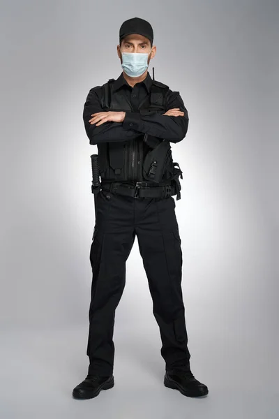 强壮的年轻警察戴着防护面具 看着工作室里的摄像头 身穿制服 双手交叉 身披灰色背景的执法警官 工作概念 — 图库照片