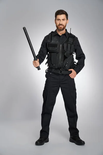 用黑色警棍对着镜头微笑英俊的警官 黑头发的警察身穿防弹衣 手持大警棍 背景灰暗 警务工作概念 — 图库照片