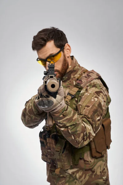 Serious bearded male sniper aiming, by pointing machine gun to camera indoors. Portrait of focused infantry man shooting with rifle, looking straight, on gray background. Concept of army, weapon.