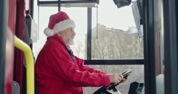 クリスマスの帽子をかぶったバス運転手のサイドビュー 夢を見る 灰色の髪とひげを持つ老人は ヘルメットに手をつないで 先を見ていて 考えています 都市生活の概念 — ストック動画