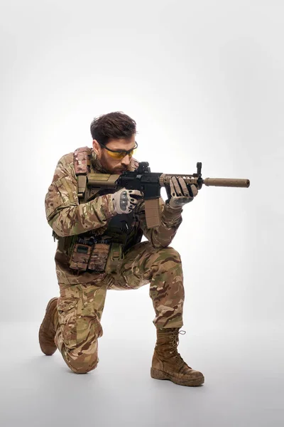 Focused special operations fighter aiming from assault rifle indoors. Side view of brave male soldier with machine gun, shooting from knee, on gray studio background. Concept of army, military force.
