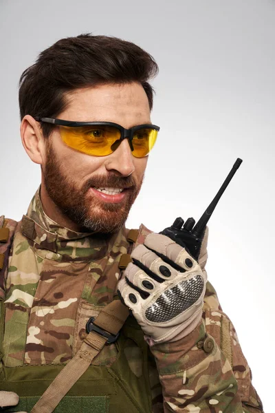 Handsome military man in yellow tactical glasses, talking on walkie-talkie. Portrait of smiling soldier in camouflage uniform, using portable radio, reporting, isolated on white. Connection concept.