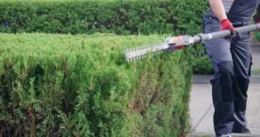 Mavi önlüklü kaslı bahçıvan ve arka bahçede elektrikli budama makinesiyle çalıları kesen eldivenler. Erkek işçilerin yavaş çekimde ve açık havada mevsimsel iş yapmalarının görüntüsü. Çalışma alanı kavramı.