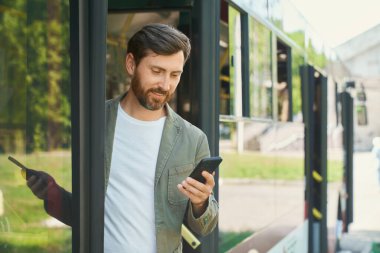 Şık bir genç adam akıllı telefon tutuyor ve şehirde otobüsten iniyor. Otobüsten inerken cep telefonunu kontrol eden sakallı adamın ön görüntüsü. Ulaşım, zamanlama kavramı.