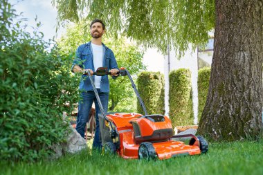 Bahçesinde elektrikli çim biçme makinesi olan mavi gömlekli yakışıklı adam. Güler yüzlü erkek bahçıvanın çim biçme makinesiyle durup, gündüzleri bahçede çalışmasının düşük açılı görüntüsü. Bahçıvanlık kavramı.