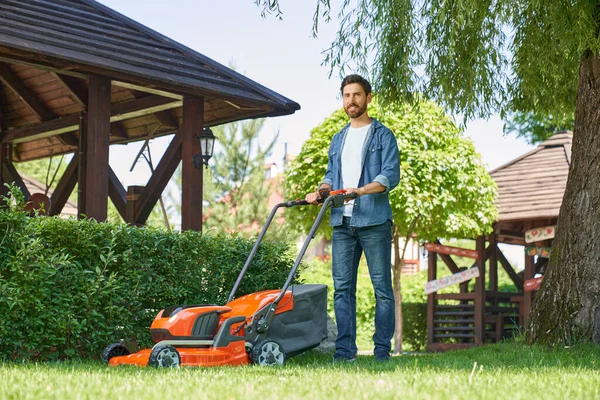 Smiling male landscaper in denim shirt trimming overgrown lawn with lawn mover at summer day. Low angle view of happy bearded guy using electric mower, while smiling at camera. Concept of gardening.