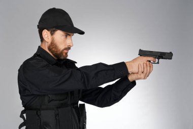 Stüdyoda tabancayla nişan alan odaklanmış sakallı polis memuru. Beyaz polisin ateş edişinin yan görüntüsü, silah tutuşu, başka tarafa bakma, gri arka planda izole edilmiş. Tehlike işi, silah kavramı.
