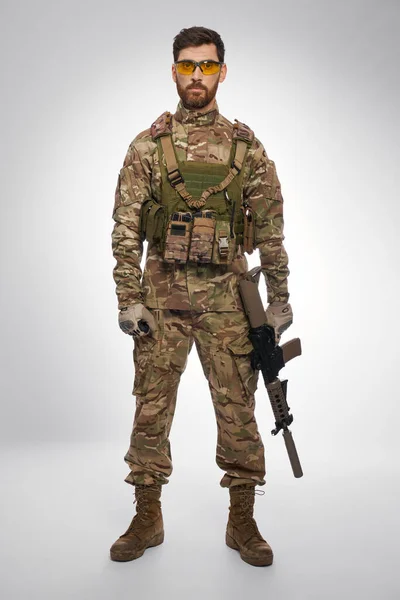 Serious male soldier in body armor holding rifle, while posing indoors. Front view of dark-haired military guy in camouflage with weapon, looking camera, with gray background. Military force concept.