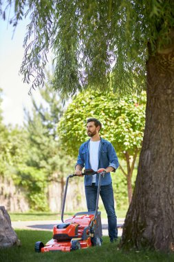 Ciddi bir erkek tamirci parkta kablosuz çim biçme makinesiyle taze çimenleri buduyor. Odaklanmış orta yaşlı bir adamın çim biçme makinesi kullanması, bahçede çalışması, mesafeye bakması. İş yeri kavramı.