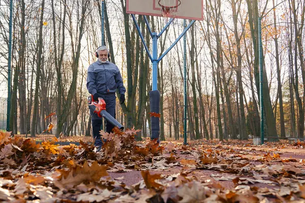Qualified maintenance worker taking care of city park area during autumn season. Low angle view of aged man in overalls removing dry leaves with leaf blower at basketball court. Seasonal work concept.