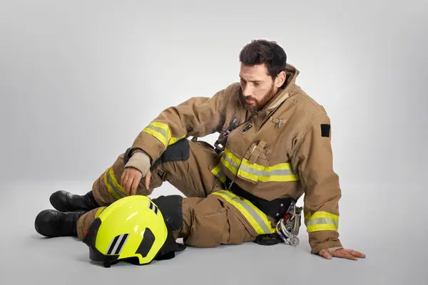 Tired male firefighter taking breath, while sitting on floor. Side view of despaired fireman taking rest after fire fighting, on gray studio background. Concept of emotions, hard work, stress.