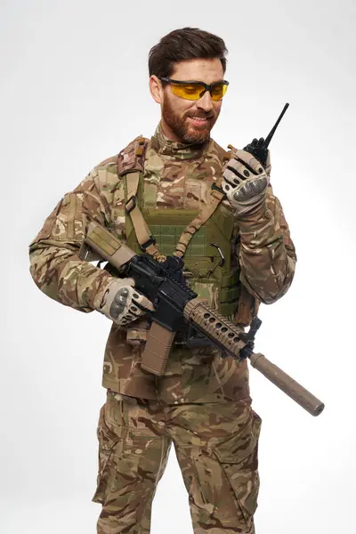 Smiling military man in tactical glasses, talking on walkie-talkie in studio. Front view of happy infantryman with assault rifle, reporting by radio, isolated on white. Army, connection concept.