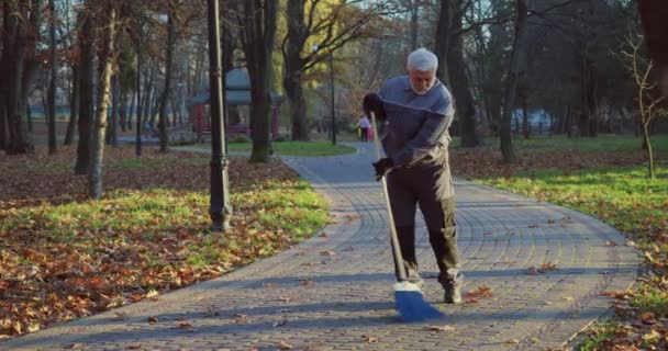 穿着工作服和手套的老人用蓝色扫帚扫地 白人看门人清除人行道上的干叶 — 图库视频影像