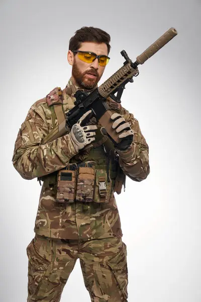 Focused male soldier in camouflage outfit recharging sniper rifle indoors. Portrait of caucasian military man in tactical gloves, looking at weapon, on gray background. War, military force concept.