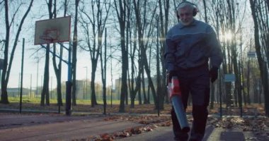 İş kıyafetleri, eldivenler ve kulaklıklar takmış, parktaki kuru yaprakları elektrikli süpürgeyle temizleyen kıdemli beyaz adam. Mevsimlik iş ve bakım kavramı.