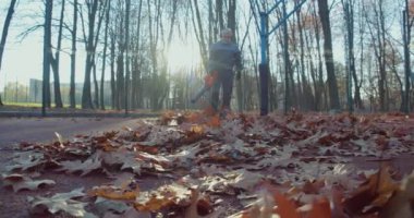 İş kıyafetleri, eldivenler ve kulaklıklar takan kıdemli bir adam şehir parkındaki kuru yaprakları kurutma makinesiyle temizliyor. Açık alanların temizleme süreci.