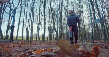 Beyaz bahçıvan, sonbahar zamanı şehir parkını temizlerken yaprak üfleyici kullanıyor. Özel tulum, güvenlik kulaklığı ve eldiven giyen erkek hademe..