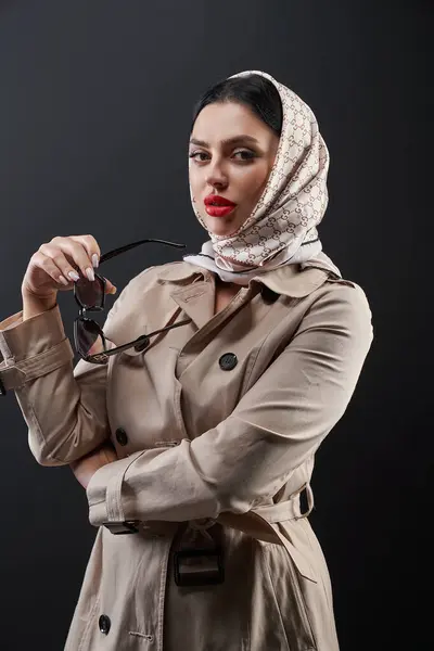 Gorgeous red lipped woman in trendy outfit looking at camera pensively. Portrait of beautiful caucasian lady in trench coat posing over dark background. Concept of modelling, fashion, style.