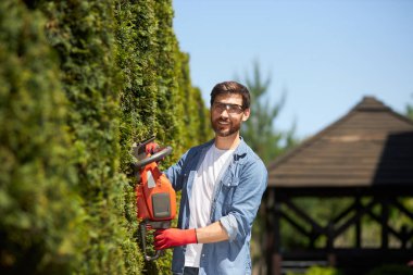 Profesyonel bahçıvan buduyor, yazın modern elektrikli budayıcı ile her zaman yeşil olan thuja çitini şekillendiriyor. Kameraya bakıp budarken gülümseyen işçinin portresi. Bahçe hizmetleri kavramı.