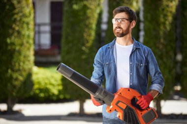 Arka bahçede modern kablosuz yaprak üfleyici taşıyan çekici sakallı erkek işçi. Gözlüklü gülümseyen bahçıvanın portresi bulanık arka planda güçlü ekipmanlar çalıştırıyor. Mevsimlik iş kavramı.