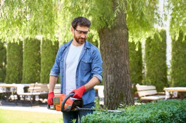 Koruyucu gözlüklü beyaz adam dışarıda çit biçme makinesiyle çalı çırpı kesiyor. Profesyonel bahçıvanın, bitkilerle ilgilenirken modern aletleri kullanması. Mevsimlik iş kavramı.