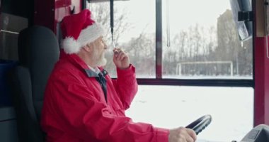Noel şapkalı otobüs şoförünün mikrofonla konuşmasının yan görüntüsü. Gri saçlı yaşlı adam dümende el ele tutuşuyor, ileriye bakıyor. Kentsel yaşam ve tatil kavramı.