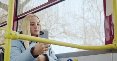 Otobüste selfie çeken sarışının yan görüntüsü. Güzel, genç bir bayan gülümsüyor, saçlarına dokunuyor, ön kameraya bakıyor, evli, mutlu, oturan yolcular. Kentsel yaşam kavramı.