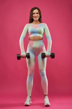 Mutlu kahverengi saçlı sporcu kadın stüdyoda dambıllarla egzersiz yapıyor. Spor yaparken halterli spor aletleri kullanan güzel bir bayan sporcunun ön görüntüsü. Pembe üzerine izole edilmiş. Spor anlayışı.