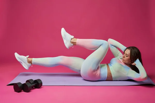Focused woman in activewear keeping hands behind head, having abdominal crunch workout. Side view of fit female doing exercise for core, lying on math against pink studio background. Sport concept.