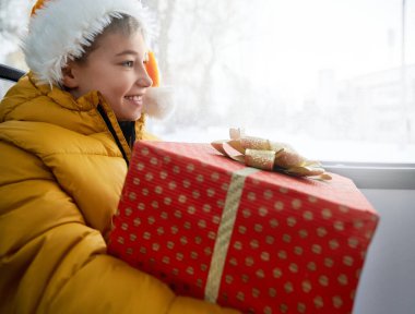 Portakal rengi Noel Baba şapkalı mutlu bir erkek çocuğu. Kırmızı noktalı kırmızı kağıt, ileriye bakıyor. Toplu taşıma aracında oturmuş gülümsüyor. Tatilde otobüsle seyahat eden çocuğa yakın durun. Mutluluk kavramı.