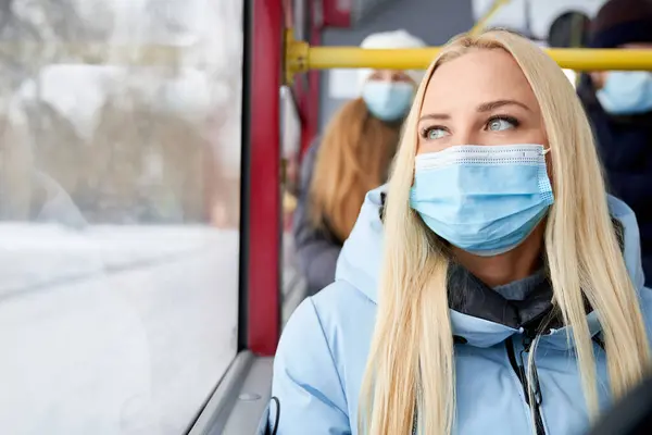 Koruyucu maske takan ve mavi ceketli güzel bir kadın otobüsün arkasında yolcularla oturuyor. Yetişkin bir kadının hareket halindeki pencerede dalgın dalgın baktığı görüntüler. Şehir hayatı kavramı.